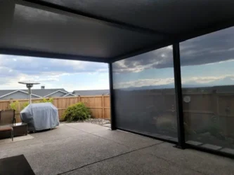 A covered patio with SkyShades retractable mesh shades providing overhead and side protection. The space has textured flooring, outdoor furniture including a covered grill, and a wooden fence enclosing the backyard. Beyond the fence, rooftops and a cloudy sky with mountains in the distance are visible.