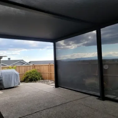 A covered patio with SkyShades retractable mesh shades providing overhead and side protection. The space has textured flooring, outdoor furniture including a covered grill, and a wooden fence enclosing the backyard. Beyond the fence, rooftops and a cloudy sky with mountains in the distance are visible.