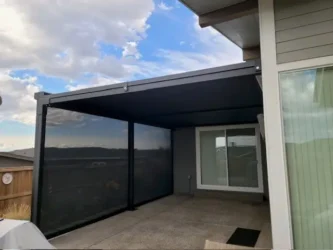 A residential backyard patio with SkyShades retractable mesh shades providing shade on the roof and two sides. The patio has a concrete floor, sliding glass doors leading into the house, and a view of neighboring rooftops and a partly cloudy sky.
