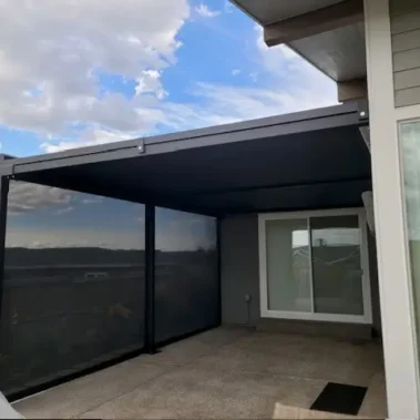 A residential backyard patio with SkyShades retractable mesh shades providing shade on the roof and two sides. The patio has a concrete floor, sliding glass doors leading into the house, and a view of neighboring rooftops and a partly cloudy sky.