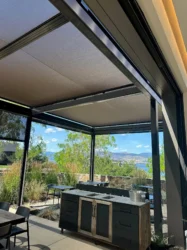 An outdoor patio with SkyShades retractable mesh shades overhead and partially lowered on the sides. The area includes tables, chairs, and a portable cabinet with a metal countertop. The background shows stone walls, plants, and a scenic view of mountains and a lake under a blue sky with clouds.