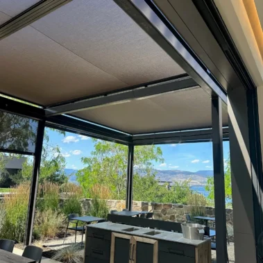An outdoor patio with SkyShades retractable mesh shades overhead and partially lowered on the sides. The area includes tables, chairs, and a portable cabinet with a metal countertop. The background shows stone walls, plants, and a scenic view of mountains and a lake under a blue sky with clouds.
