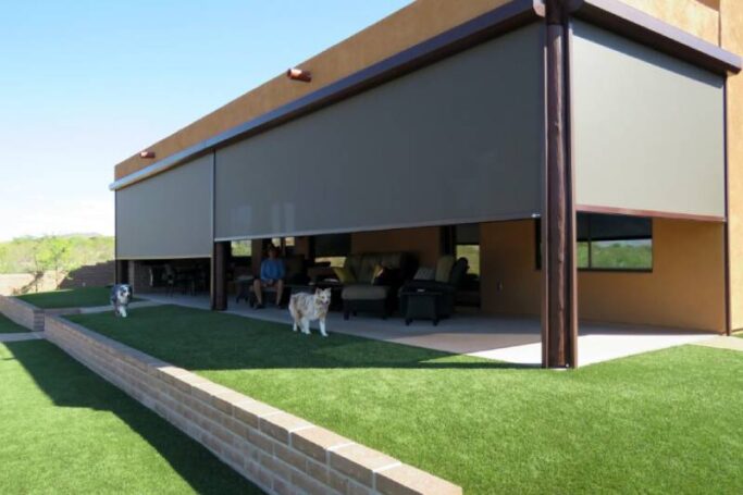 Solar screen halfway down across a patio opening with the homeowners relaxing inside and their two dogs run across the grassy yard.