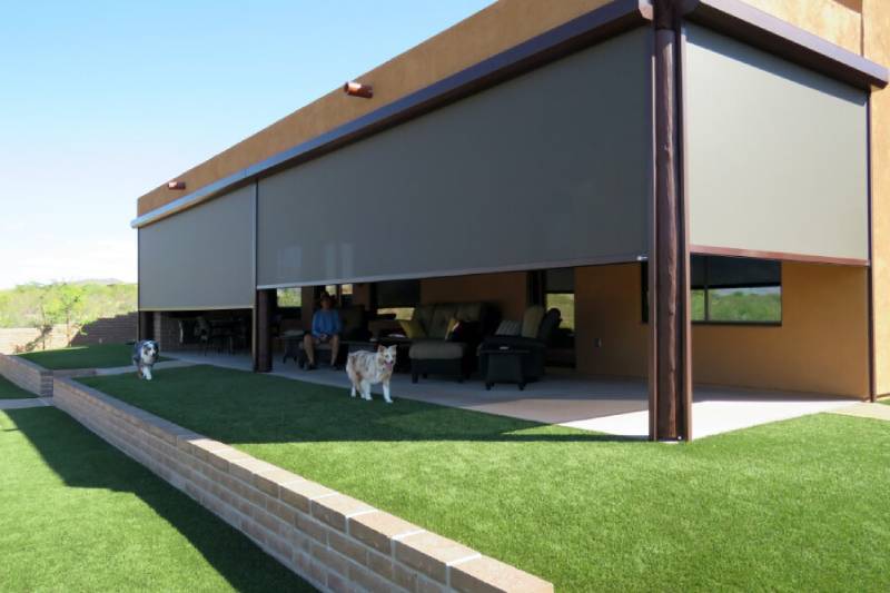 Solar screen halfway down across a patio opening with the homeowners relaxing inside and their two dogs run across the grassy yard.