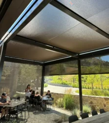 An outdoor dining area with gravel flooring, modern black tables and chairs, and retractable mesh SkyShades providing overhead and side shade. A group of people sits at one table while a woman stands nearby. The background shows a stone wall, greenery, and a winding road under a sunny sky.