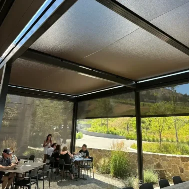 An outdoor dining area with gravel flooring, modern black tables and chairs, and retractable mesh SkyShades providing overhead and side shade. A group of people sits at one table while a woman stands nearby. The background shows a stone wall, greenery, and a winding road under a sunny sky.