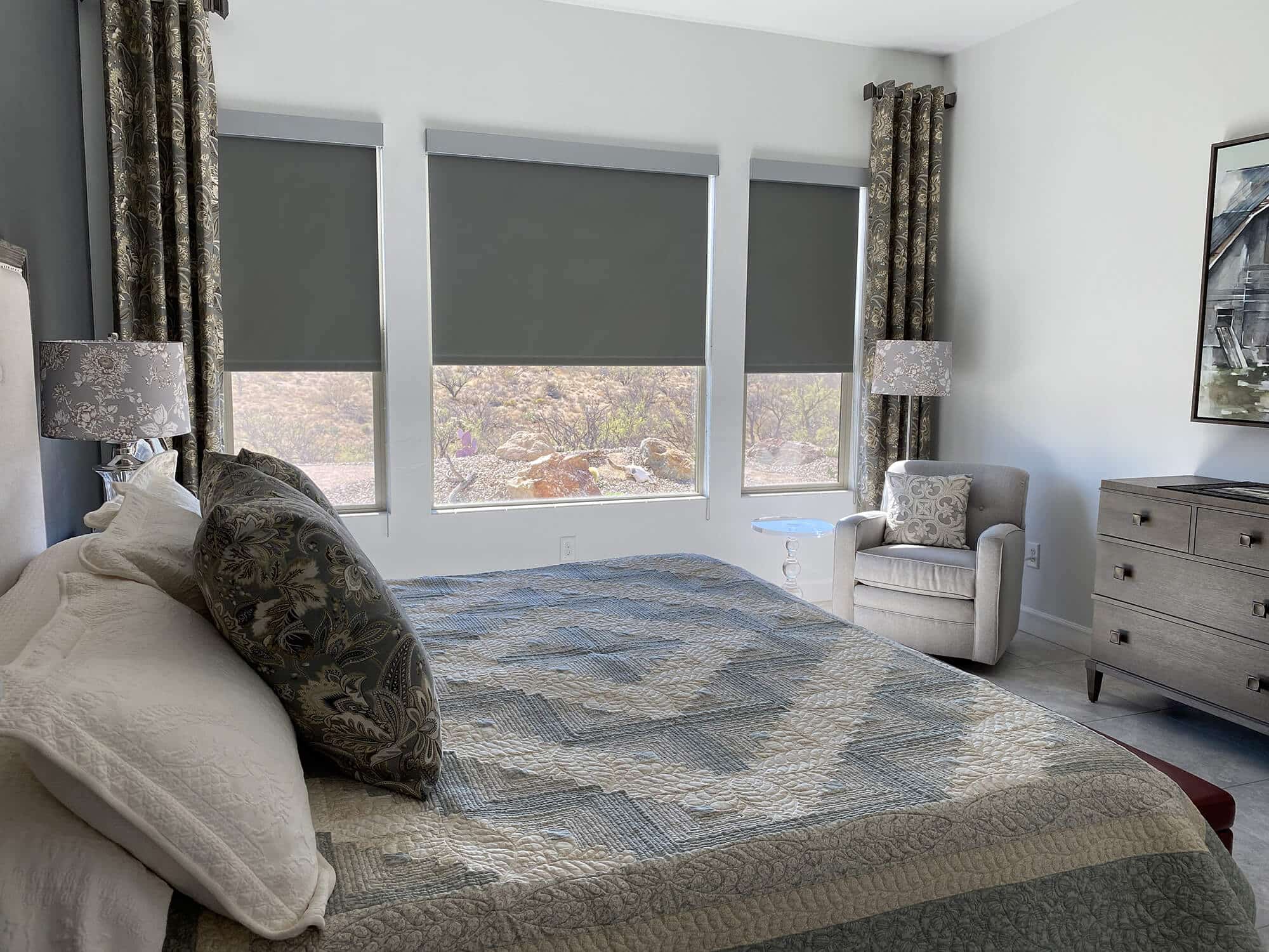 Modern bedroom with patterned bedding, armchair, and large windows featuring gray automated roller blinds.