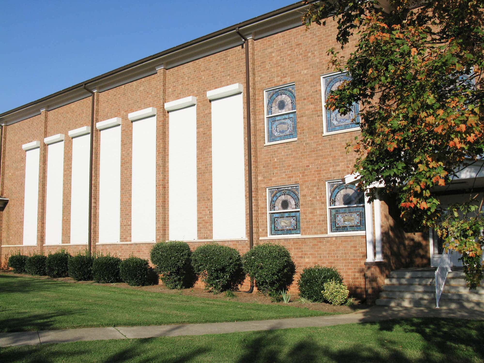Brick building with large windows, surrounded by green shrubs and trees, under a clear blue sky.