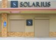 Closed storefront with Solarius sign, beige roller shutters, and brick accents on a sunny day.