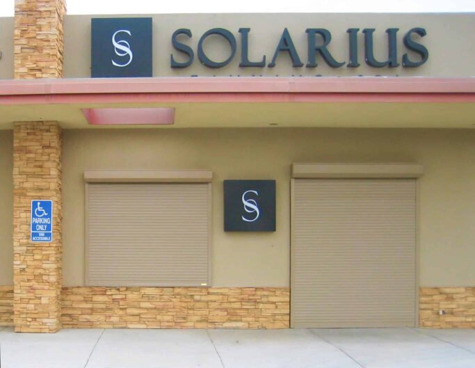 commercial-shutters-solarius Closed storefront with Solarius sign, beige roller shutters, and brick accents on a sunny day.